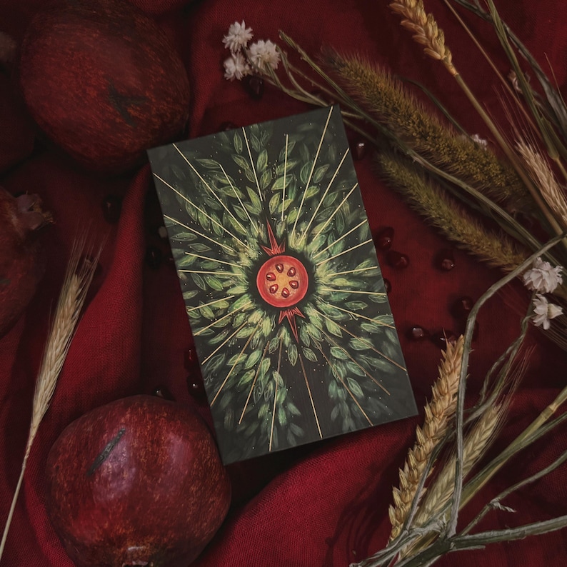 May include: A rectangular deck of cards featuring a dark green background with a central illustration of a sliced pomegranate surrounded by radiating golden lines and green leaves. The artwork is detailed with a symmetrical botanical pattern.