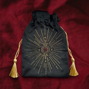 Pode incluir: Uma bolsa de veludo preto com um design de estrela de explosão bordado em ouro. A bolsa tem um fecho de cordão e borlas douradas.