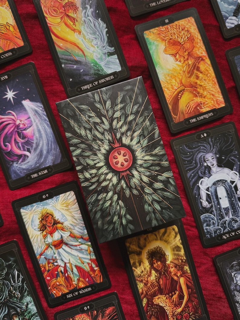 May include: A deck of tarot cards featuring a central box with a pomegranate illustration surrounded by green leaves and radiating lines. Surrounding cards display colourful, detailed illustrations including The Empress, The Star, and Six of Wands.