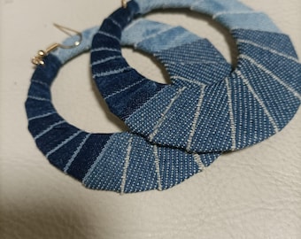 May include: A pair of denim hoop earrings. The earrings are made of strips of blue denim fabric wrapped around a circular frame. The denim has a gradient effect, transitioning from dark to light blue. The earrings have gold-coloured ear wires.