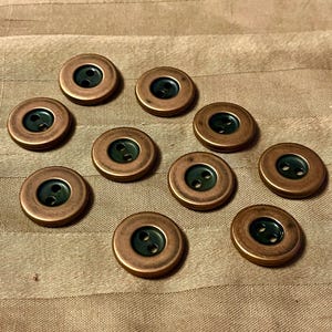 Peut inclure: Dix boutons de couleur cuivre avec un centre vert foncé et deux trous. Les boutons sont ronds avec un bord légèrement surélevé. Ils sont disposés sur un tissu beige rayé.