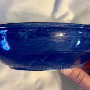 May include: A dark blue ceramic bowl with a shiny surface. The bowl has a rounded form with a gently sloping rim and a textured exterior. The bowl is held by a hand.