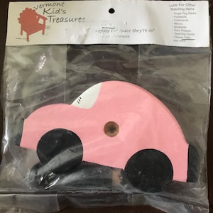 Könnte beinhalten: Ein rosa Holz-Schubladengriff in Form eines Autos mit schwarzen Rädern und einem braunen Knopf. Das Auto ist in einer transparenten Plastiktüte verpackt. Auf der Verpackung steht "Vermont Kid's Treasures".