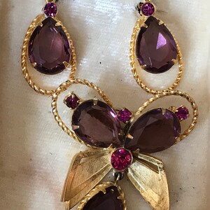 May include: A vintage jewelry set featuring a gold-toned necklace and earrings. The set includes teardrop-shaped purple gemstones and smaller pink accents. The necklace has a bow design with leaf details, and the earrings have a circular top.