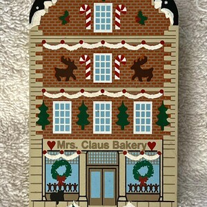 May include: A wooden Christmas decoration in the shape of a bakery. The building features a brick facade with windows, candy canes, and reindeer. The sign reads "Mrs. Claus Bakery" with a heart symbol.