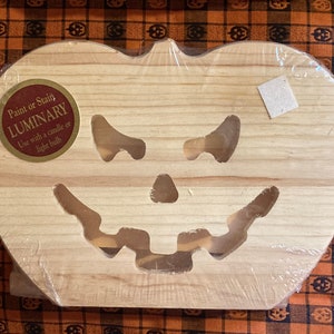 Op de afbeelding: Een houten pompoenvormige lantaarn met een uitgesneden gezicht, ontworpen voor gebruik met een kaars of gloeilamp. De lantaarn is gemaakt van lichtgekleurd hout en is verpakt in doorzichtig plastic. Het product heeft een sticker met de tekst "Paint or Stain LUMNIARY".