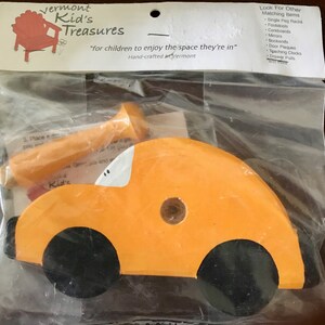 May include: An orange wooden toy car with black wheels and white accents, packaged in a clear plastic bag. The package includes a wooden peg and the text "Vermont Kid's Treasures".