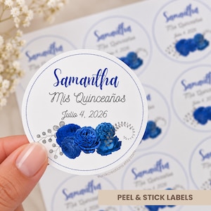 Può includere: Un adesivo circolare bianco con il testo blu Samantha, Mis Quinceaños e la data Julio 4, 2026. Il design presenta un gruppo di rose blu intenso e fogliame decorativo argentato nella parte inferiore, il tutto racchiuso da un bordo punteggiato.