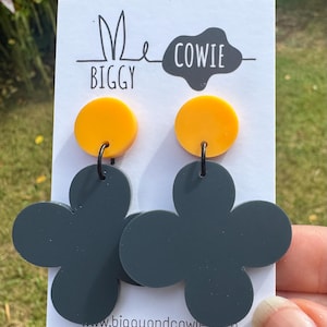 May include: A pair of earrings with a yellow disc top and a dark grey flower-shaped bottom. The earrings are connected with a black hook. The brand name "Me Biggy Cowie" is printed on the white card.