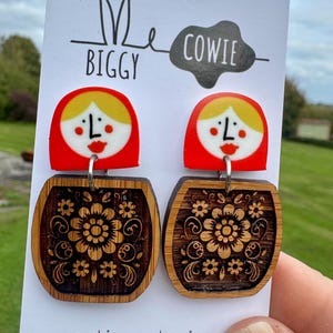 May include: Earrings with a Matryoshka doll design. The top is red with a yellow head and a painted face. The bottom is a wooden barrel shape with a floral design. The earrings hang from a white card with the text "Me Biggy Cowie". The website address is also visible.