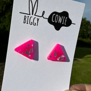 May include: A pair of pink triangle shaped earrings with silver glitter embedded in the resin. The earrings are displayed on a white card with the text "Me Biggy Cowie" and "www.biggyandcowie.com".