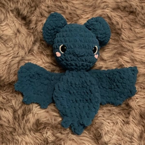 May include: A teal blue crocheted bat plush toy with black eyes and pink cheeks. The toy is lying on a brown fuzzy surface.