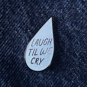 May include: A blue enamel pin shaped like a teardrop with the text "LAUGH TIL WE CRY" in white.