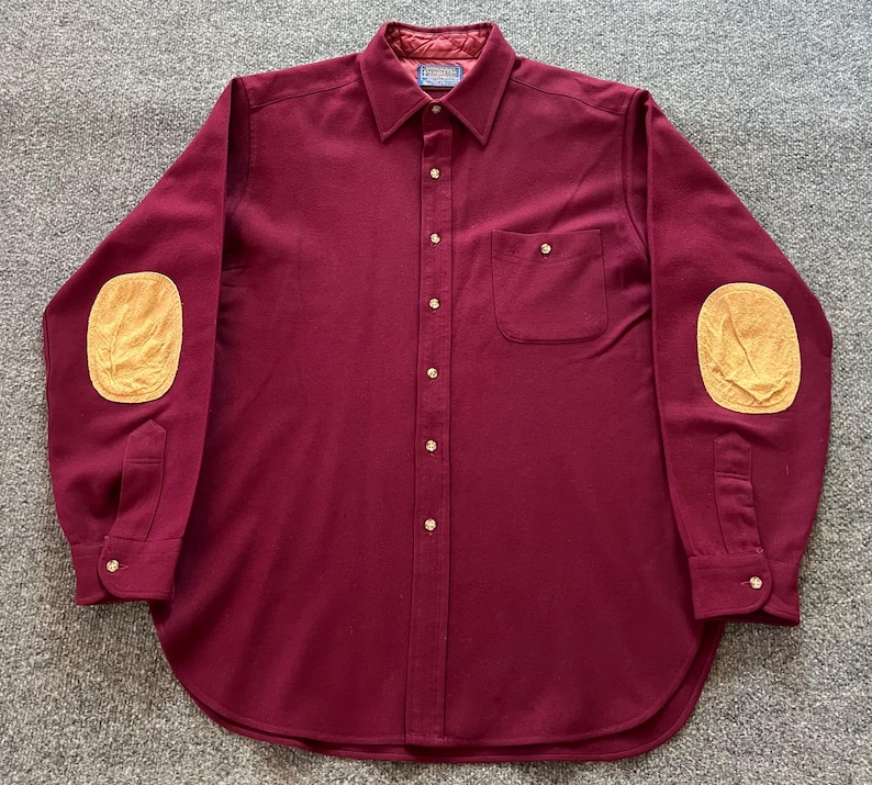 May include: A long-sleeved button-down shirt in a deep burgundy color featuring a pointed collar, a single chest pocket, and contrasting tan-colored elbow patches. The shirt has a front button placket and buttoned cuffs with a quilted inner collar.