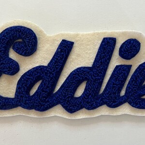 May include: A custom name patch featuring the name Eddie embroidered in a dark blue, textured, cursive script font. The lettering is stitched onto a cream-colored, felt-like fabric backing that follows the contour of the name.