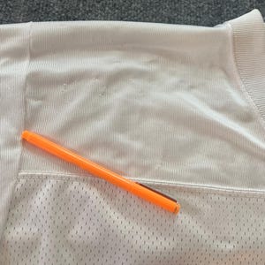 Op de afbeelding: Close-up van een wit mesh shirt met een oranje pen erop. Het shirt heeft een V-hals en een getextureerde, ademende stof. De pen is feloranje met een zilveren clip.