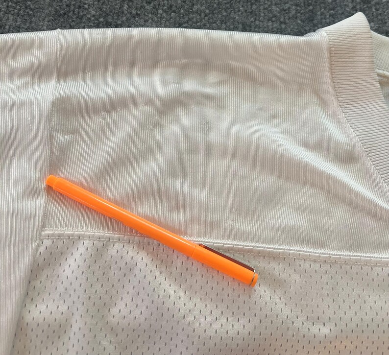 Op de afbeelding: Close-up van een wit mesh shirt met een oranje pen erop. Het shirt heeft een V-hals en een getextureerde, ademende stof. De pen is feloranje met een zilveren clip.