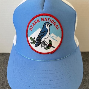 May include: A light blue trucker hat with a white mesh back and a curved brim. The front features a circular patch with the text "OZARK NATIONAL" around a blue bird illustration.