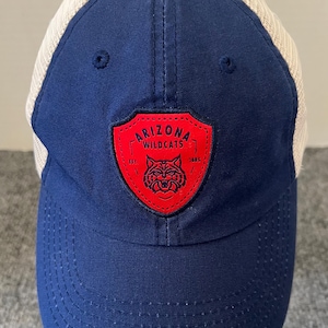 May include: A navy blue baseball cap with a mesh back. The front of the cap features a red patch with the words "ARIZONA WILDCATS" and a wildcat logo. The cap is a classic style with a curved brim.