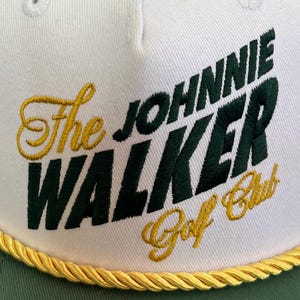 May include: White and green baseball cap with the text "The JOHNNIE WALKER Golf Club" in green and gold embroidery. The cap has a gold rope trim along the brim.