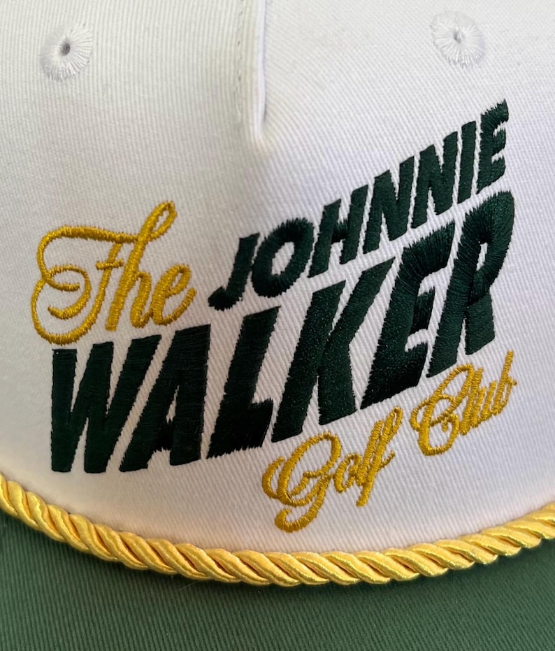 May include: White and green baseball cap with the text "The JOHNNIE WALKER Golf Club" in green and gold embroidery. The cap has a gold rope trim along the brim.