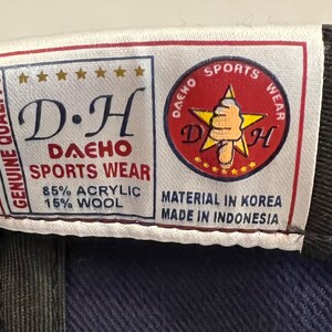 Pu&ograve; includere: Un'etichetta bianca con testo nero che recita "GENUINE QUALITY D.H DAEHO SPORTS WEAR 85% ACRYLIC 15% WOOL" e una stella rossa con contorno bianco e il testo "DAEHO SPORTS WEAR D.H" all'interno. L'etichetta riporta anche il testo "MATERIAL IN KOREA MADE IN INDONESIA".