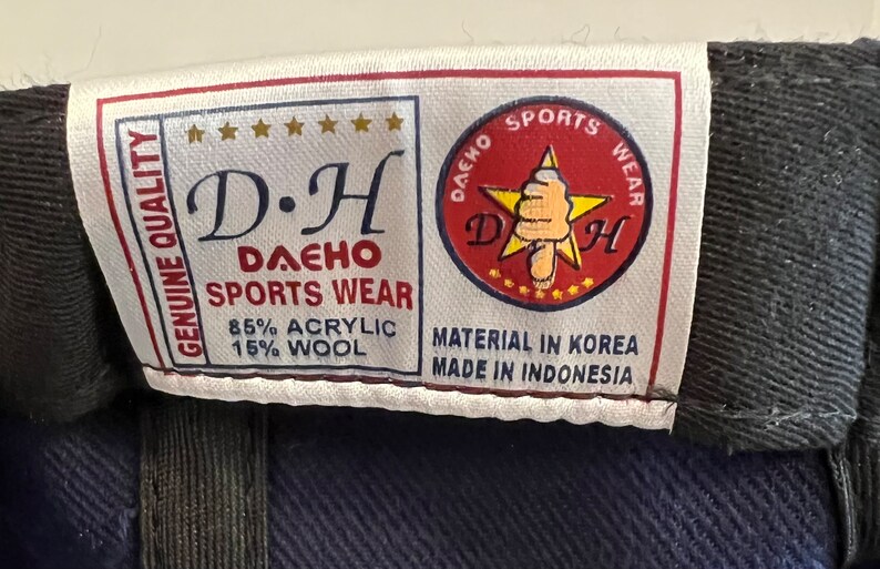 Pu&ograve; includere: Un'etichetta bianca con testo nero che recita "GENUINE QUALITY D.H DAEHO SPORTS WEAR 85% ACRYLIC 15% WOOL" e una stella rossa con contorno bianco e il testo "DAEHO SPORTS WEAR D.H" all'interno. L'etichetta riporta anche il testo "MATERIAL IN KOREA MADE IN INDONESIA".