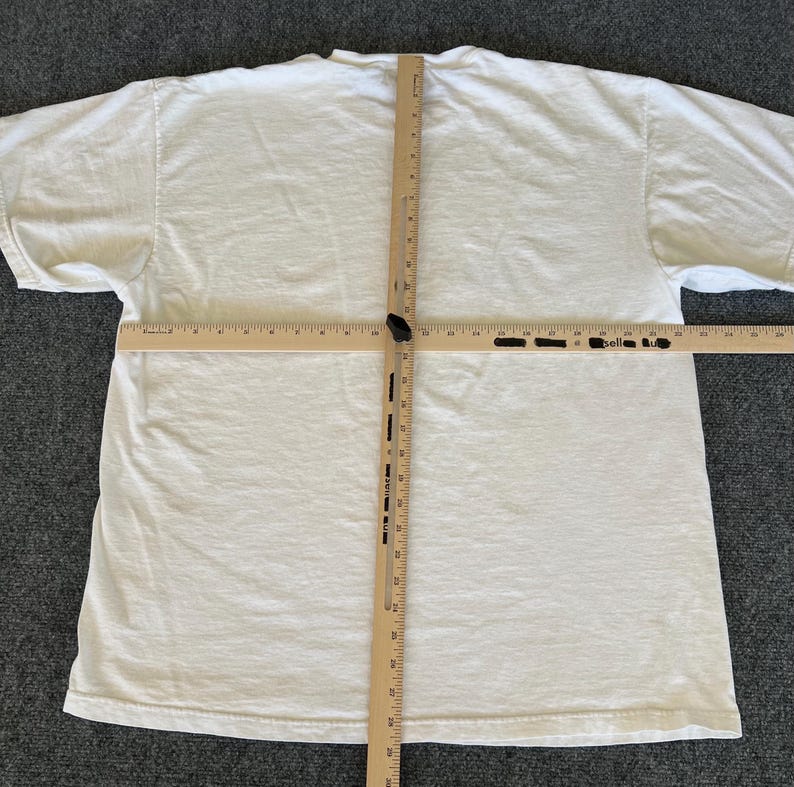 May include: A plain white t-shirt, laid flat on a gray surface, with a wooden ruler placed across the shirt to measure its dimensions. The shirt is made of a soft, likely cotton material.