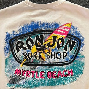 Peut inclure: Un sweat-shirt couleur cr&egrave;me avec une impression graphique repr&eacute;sentant un surfeur sur une planche jaune et rose. Le texte indique RON JON SURF SHOP en noir et MYRTLE BEACH en rose, entour&eacute; d'un motif de vagues bleu et sarcelle.