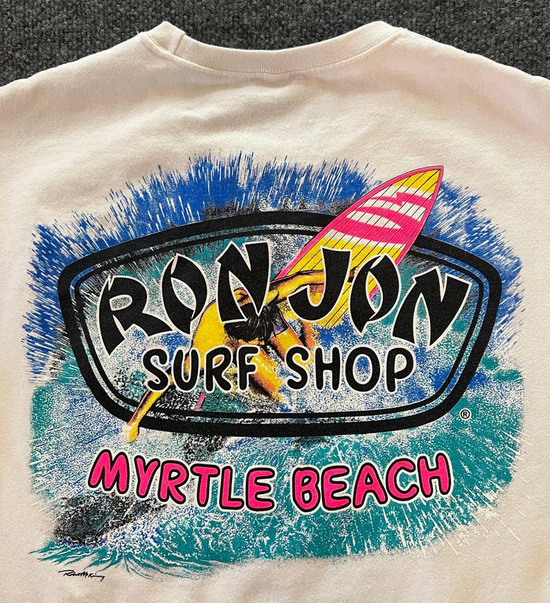 Peut inclure: Un sweat-shirt couleur cr&egrave;me avec une impression graphique repr&eacute;sentant un surfeur sur une planche jaune et rose. Le texte indique RON JON SURF SHOP en noir et MYRTLE BEACH en rose, entour&eacute; d'un motif de vagues bleu et sarcelle.