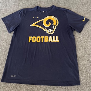 Op de afbeelding: Marineblauw T-shirt met korte mouwen met een groot goud en wit ramshoofdlogo en het woord "FOOTBALL" in gouden blokletters. Voorzien van het Nike-logo in goud. Gemaakt van Dri-FIT materiaal.