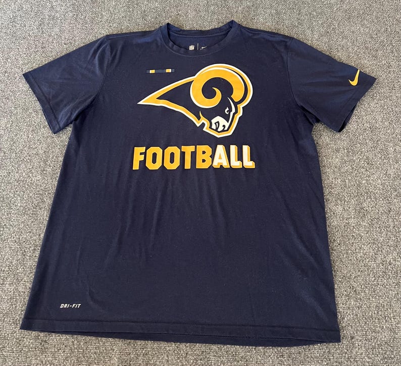 Op de afbeelding: Marineblauw T-shirt met korte mouwen met een groot goud en wit ramshoofdlogo en het woord "FOOTBALL" in gouden blokletters. Voorzien van het Nike-logo in goud. Gemaakt van Dri-FIT materiaal.