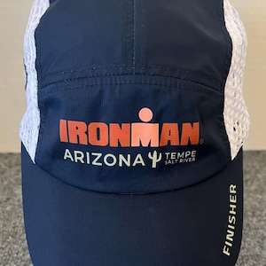 May include: A navy blue athletic cap with white mesh side panels. The front features the orange and white text IRONMAN ARIZONA TEMPE SALT RIVER with a cactus icon. The word FINISHER is printed in white vertically along the side of the curved brim.