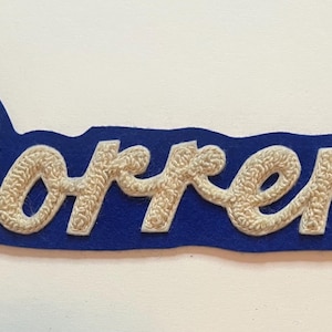 May include: A blue felt patch featuring the name Dorrena embroidered in white script lettering. The white embroidery has a textured, raised appearance, and the blue felt backing follows the contour of the cursive text in a long, horizontal shape.