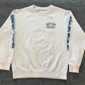 Peut inclure: Un sweat-shirt blanc &agrave; manches longues et col rond avec un petit logo bleu et noir sur la poitrine gauche indiquant Ron Jon Surf Shop Myrtle Beach. Les manches pr&eacute;sentent un imprim&eacute; graphique vertical bleu et noir le long de chaque bras.