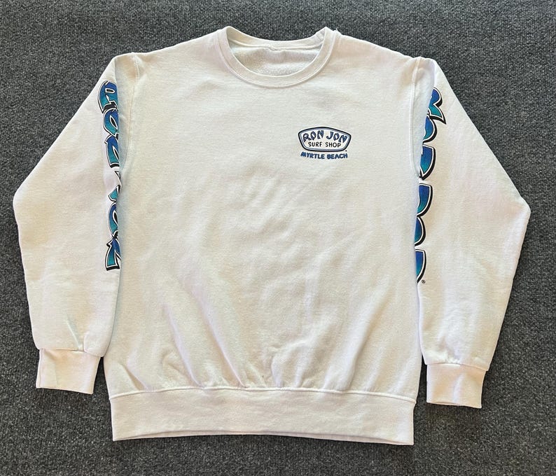 Peut inclure: Un sweat-shirt blanc &agrave; manches longues et col rond avec un petit logo bleu et noir sur la poitrine gauche indiquant Ron Jon Surf Shop Myrtle Beach. Les manches pr&eacute;sentent un imprim&eacute; graphique vertical bleu et noir le long de chaque bras.