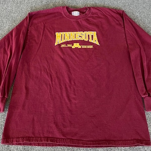 May include: A maroon long-sleeve shirt with the word "MINNESOTA" in gold lettering across the chest. Below the word "MINNESOTA" is the text "EST. 1891" and the letter "M" with the words "GOLDEN GOPHERS". The shirt has a crew neck and ribbed cuffs.