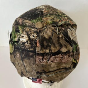May include: A camouflage baseball cap with a brown button on top. The cap features a pattern of green, brown, and tan, with an American flag tag. The cap is designed for outdoor activities.
