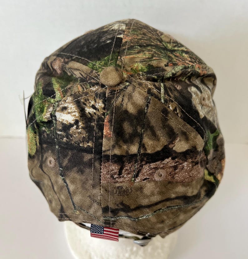 May include: A camouflage baseball cap with a brown button on top. The cap features a pattern of green, brown, and tan, with an American flag tag. The cap is designed for outdoor activities.