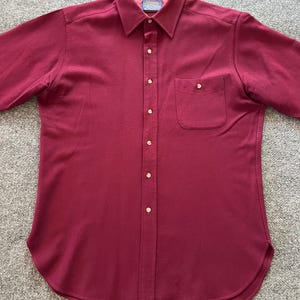 May include: A solid burgundy button-down shirt featuring a pointed collar, a single chest pocket with a button closure, and a quilted interior lining at the collar. The shirt has a front button placket with light-colored buttons and a curved hem.