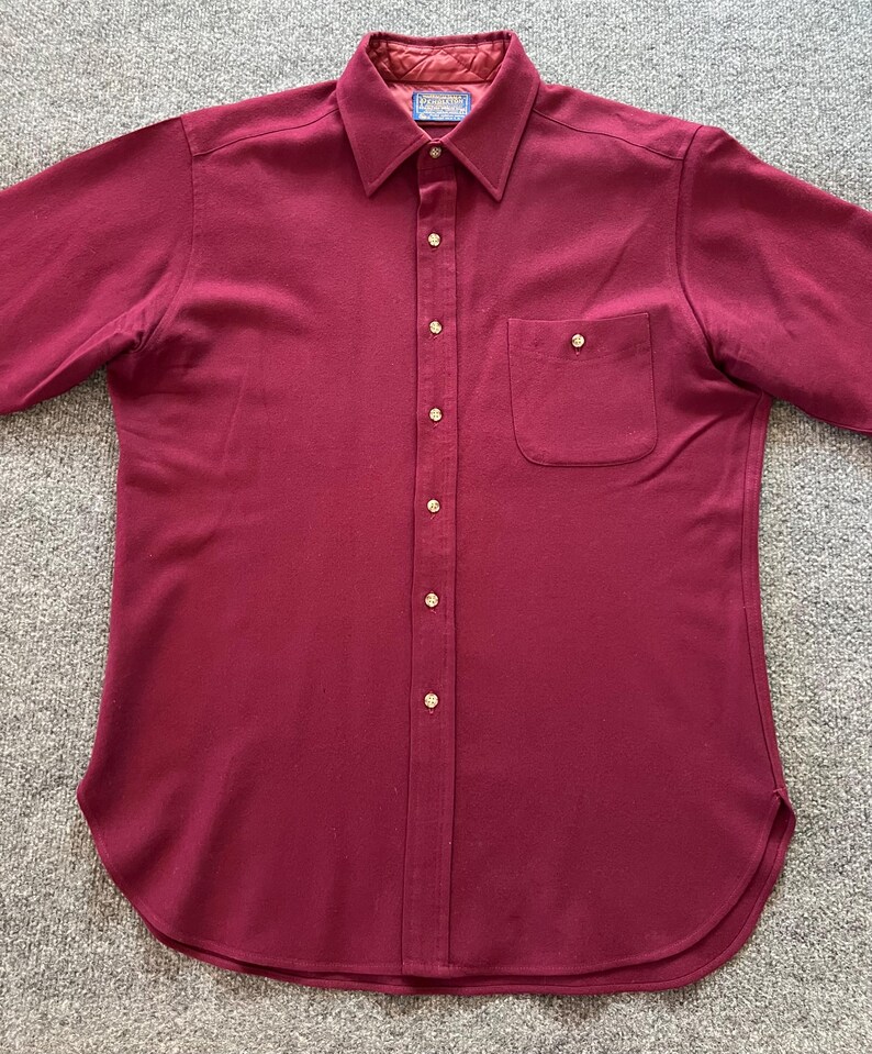 May include: A solid burgundy button-down shirt featuring a pointed collar, a single chest pocket with a button closure, and a quilted interior lining at the collar. The shirt has a front button placket with light-colored buttons and a curved hem.