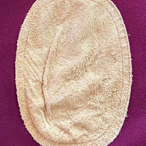 May include: An oval-shaped beige suede elbow patch with a textured, fuzzy surface and a visible stitched border along the edge, positioned against a solid deep purple fabric background.