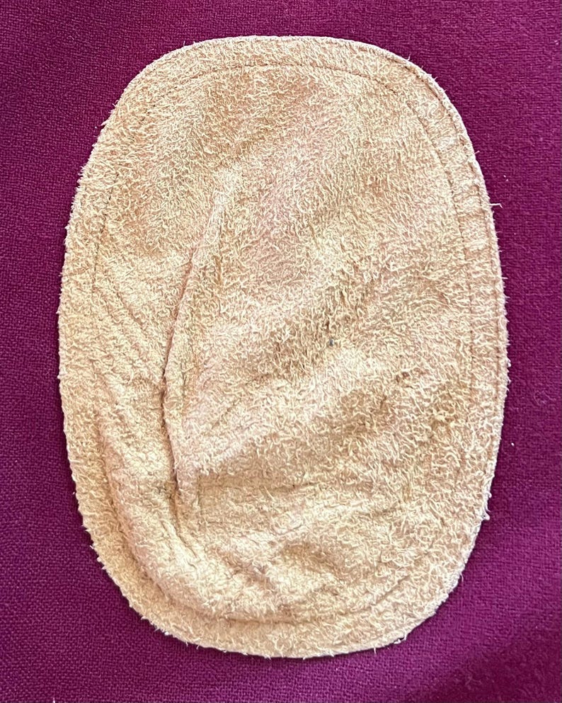 May include: An oval-shaped beige suede elbow patch with a textured, fuzzy surface and a visible stitched border along the edge, positioned against a solid deep purple fabric background.