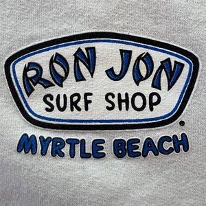 Peut inclure: Un tissu gris clair pr&eacute;sentant un logo s&eacute;rigraphi&eacute; pour Ron Jon Surf Shop. Le logo est entour&eacute; d'une forme aux contours noirs et bleus avec le texte RON JON SURF SHOP en noir et bleu, et les mots MYRTLE BEACH en bleu sous la forme.
