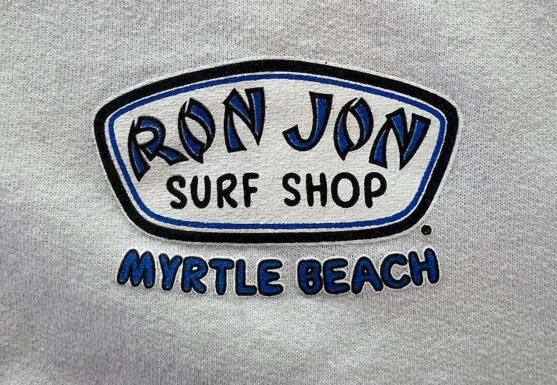 Peut inclure: Un tissu gris clair pr&eacute;sentant un logo s&eacute;rigraphi&eacute; pour Ron Jon Surf Shop. Le logo est entour&eacute; d'une forme aux contours noirs et bleus avec le texte RON JON SURF SHOP en noir et bleu, et les mots MYRTLE BEACH en bleu sous la forme.