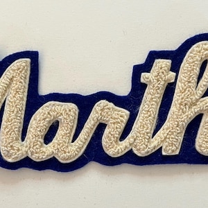 May include: A script-style name patch featuring the name Martha in raised, textured off-white embroidery. The lettering is outlined and backed by a solid, deep blue felt material, creating a bold contrast between the light text and dark border.