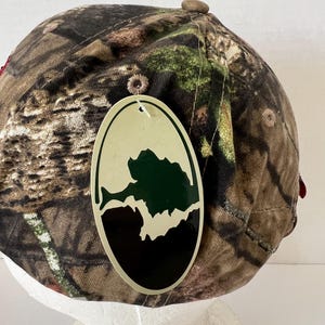 May include: A camouflage baseball cap with a brown button on top and a tag with a green and black graphic. The cap features a pattern of brown, green, and tan, typical of hunting apparel.