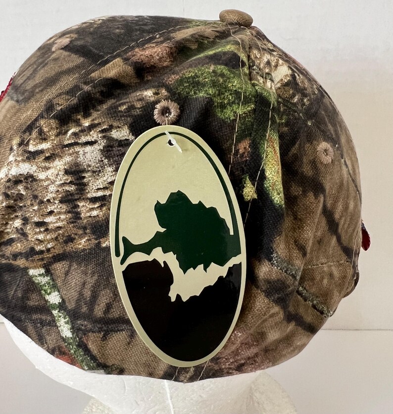 May include: A camouflage baseball cap with a brown button on top and a tag with a green and black graphic. The cap features a pattern of brown, green, and tan, typical of hunting apparel.