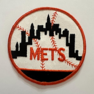 May include: A circular embroidered patch featuring a white background with a black city skyline silhouette. The design includes orange baseball stitching patterns and the word METS in orange thread, all enclosed by a thick orange border.