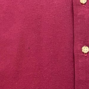 May include: A deep magenta or maroon colored shirt featuring a vertical placket with two visible light-colored, four-hole buttons. The fabric has a textured, slightly fuzzy appearance with a small, dark pinhole imperfection visible on the left side.